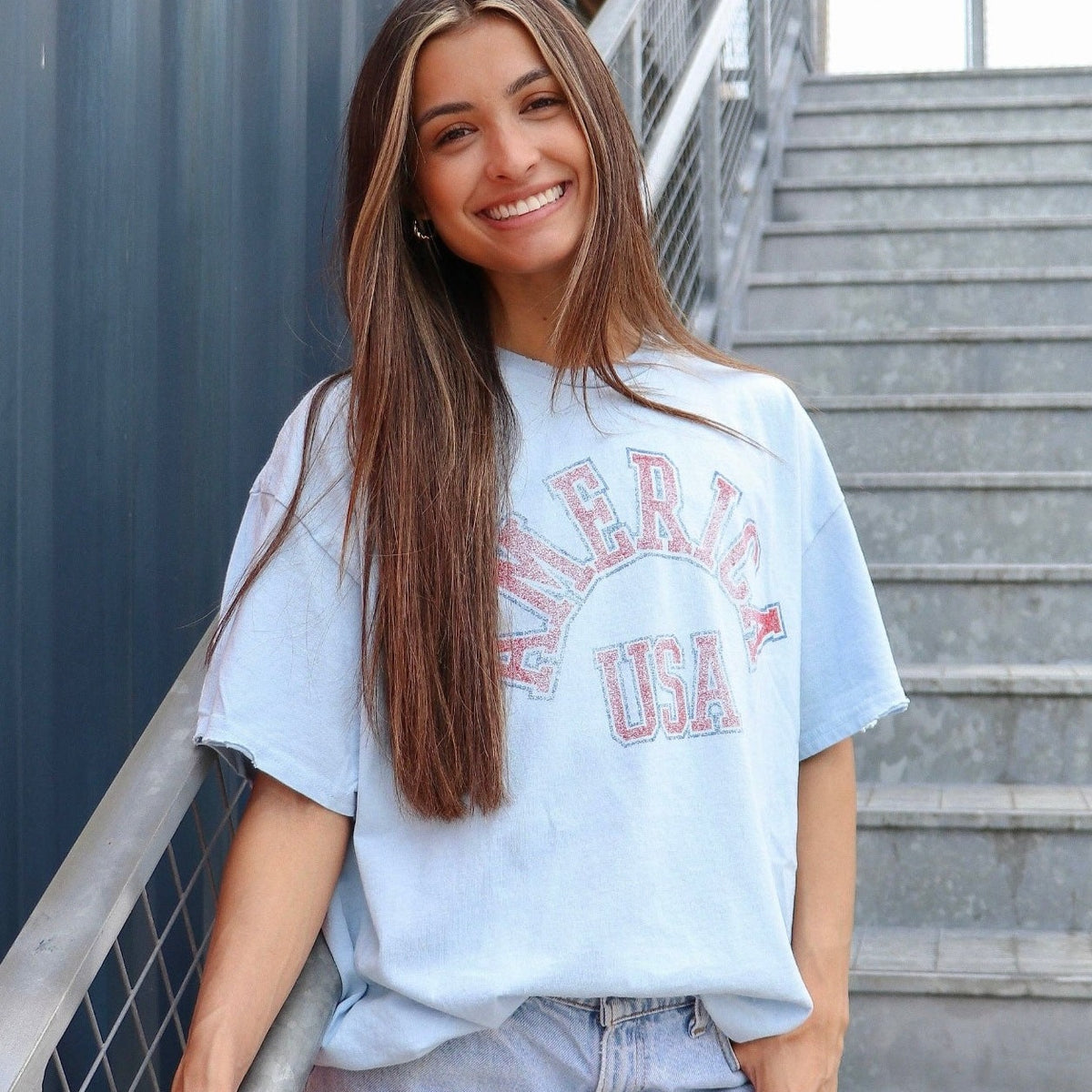 Young female standing on stairs wearing oversized white graphic t-shirt that says "America USA" in blue and red letters